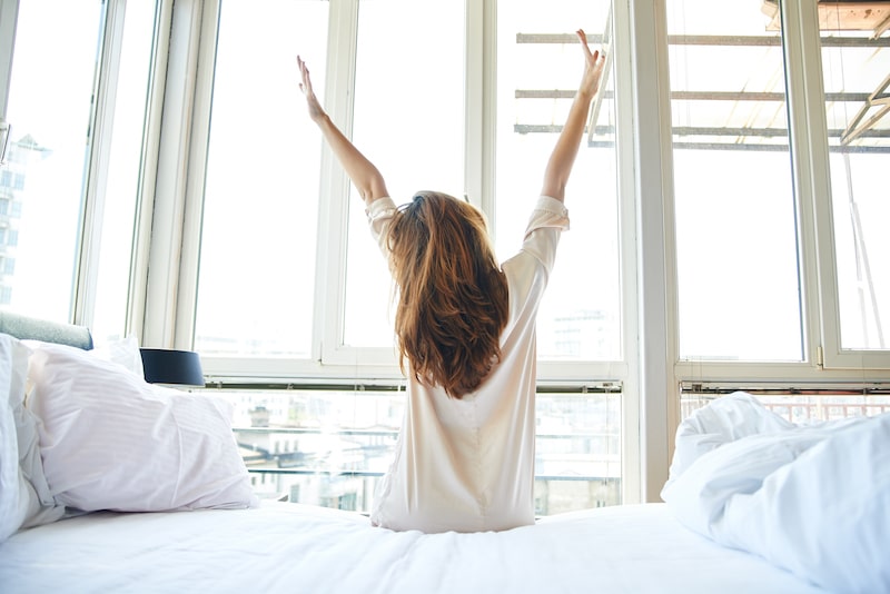 Young woman stretching her back in bed looking out over the sunrise